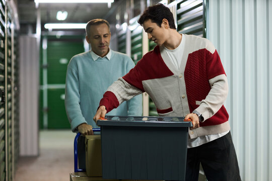 Caucasian middle aged man and Caucasian young adult man moving plastic storage box and cardboard boxes in self storage facility, both focused on organizing belongings and working together