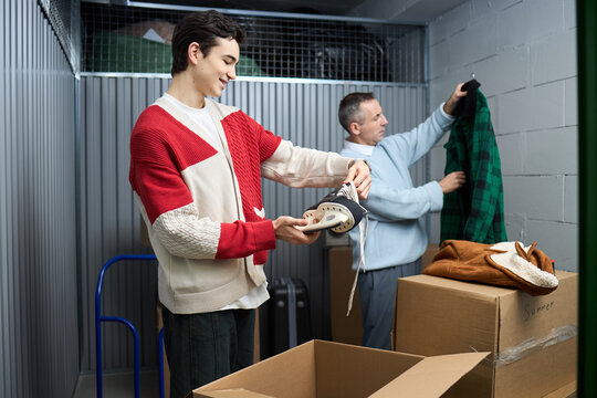 Young adult Caucasian man unpacking ice skates from cardboard box while middle aged Caucasian man sorting green plaid shirt in storage unit, both standing near open boxes