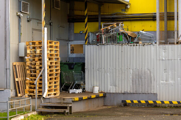 Industrial loading area shows stacked wooden pallets near metal steps and wall. Shopping carts and scrap items piled beside fenced platform.