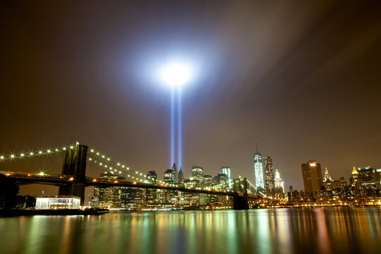 View of two beams of light pierce the night sky above the city skyline and the iconic Brooklyn Bridge, reflecting on the water, New York, New York, United States.