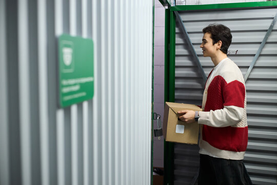 Young adult Caucasian man carrying cardboard box into storage unit, standing sideways and smiling, holding package with both hands, metal walls and open door visible in background