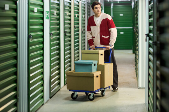 Young adult Caucasian man pushing cart loaded with cardboard boxes through corridor of self storage facility, looking forward while moving belongings, surrounded by green storage unit doors
