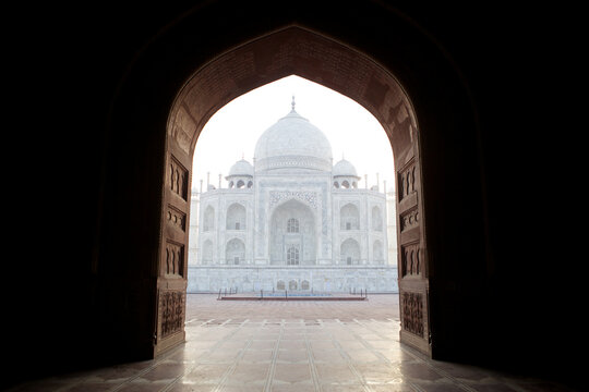 View of the Taj Mahal's pristine white marble shimmers ethereally through the dark, ornate archway, a timeless monument framed in shadow, Agra, Uttar Pradesh, India.