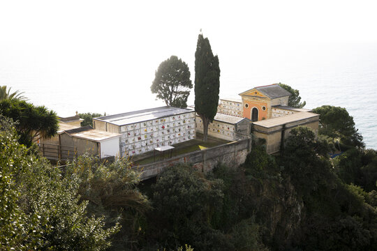 View of a serene cemetery perched atop a cliffside, overlooking the azure sea under a hazy sky, a poignant blend of life's end and nature's beauty, Manarola, Liguria, Italy.