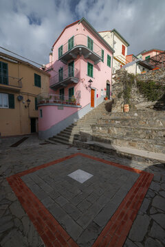 View of pastel buildings rise above a patterned square, framed by stone steps and a sky brushed with clouds, in Manarola, Liguria, Italy.