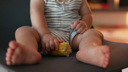 Obraz premium Toddler sits with legs outstretched, holding corn cob while seated on black chair, showcasing relaxed grip and foot positioning in casual indoor space