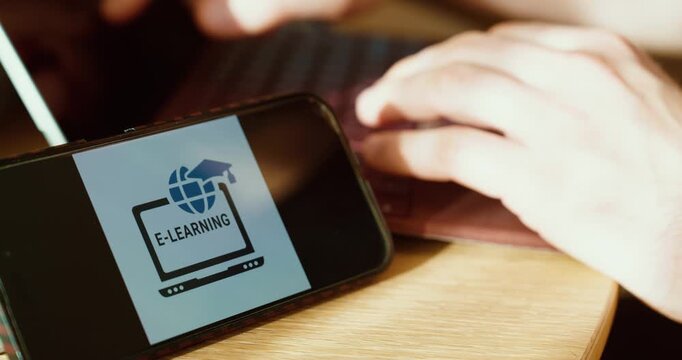 Close-up of a smartphone displaying an e-learning interface while hands type on a laptop at a desk, showing modern online training and remote education. Ideal for corporate learning, upskilling, compl