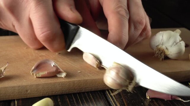 Close-up of hands slicing garlic cloves on a wooden cutting board with a sharp knife, showcasing the preparation process in a kitchen setting