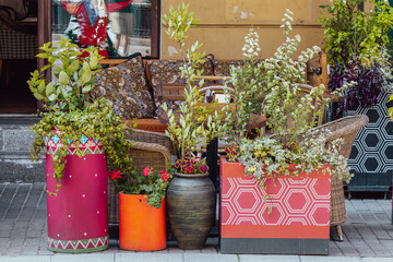 Wicker chairs and table sit behind large planters with leafy plants and flowers on sidewalk. Patterned cushions and storefront window visible.
