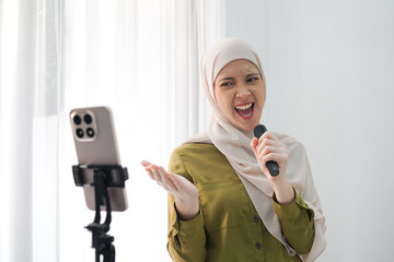 A woman wearing a hijab sings into a handheld microphone while filming with a smartphone on a tripod, creating a lively home-performance scene. © Hardollin