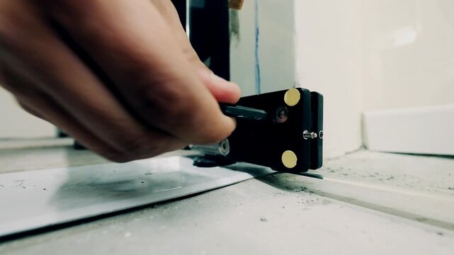 The camera is placed on the ground, a glass door hinge is really close to the camera and a workers hand enters the frame with an Allen key, starting to tighten the bolt. a very shallow depth of field 