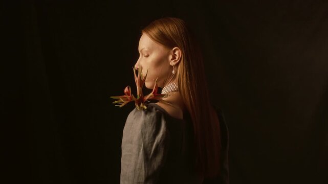 Rear view of young woman with long red hair wearing pearl necklace and earrings turning to camera while posing with hippeastrum sumatra flower in hands against dark backdrop in dim studio