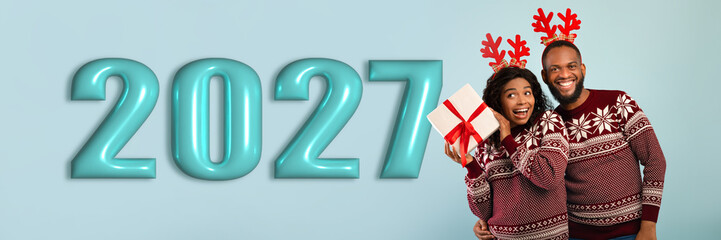 Obraz premium A couple stands together, smiling and holding a gift. They wear matching holiday sweaters and have festive reindeer antlers. The backdrop shows the year 2027 in large numbers.