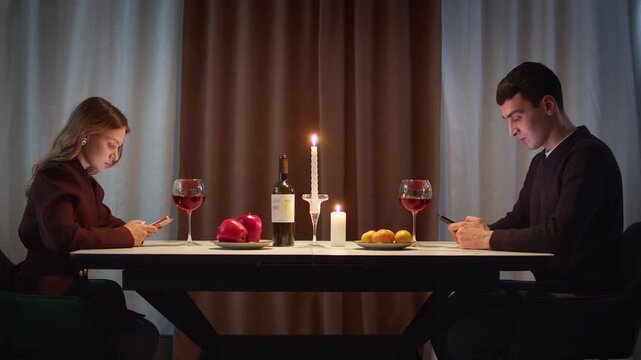 Couple with cell mobile phone addiction having evening wine dinner together. People celebrating event. Awkward ignore romantic first date problem at restaurant or home. Holiday candle light atmosphere