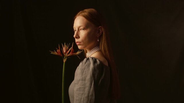 Profile view of young woman with long red hair wearing pearl earrings and necklace holding hippeastrum sumatra flower then turning to camera against dark backdrop in low key lit studio