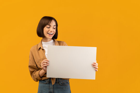 Wow. Excited woman holding white empty board for advertisement and looking down at big blank placard with open mouth, posing over yellow background, empty space for text