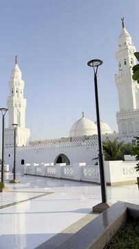 A serene exterior shot of the historic Masjid al-Qiblatayn in Medina, featuring its iconic white minarets and domes framed by vibrant yellow flowers and lush green leaves under a clear blue sky.