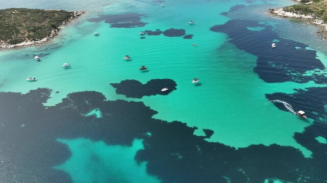Aerial drone view of boats floating over crystal clear turquoise Mediterranean water with dark underwater reef patterns, Greece. Top-down seascape showing shallow lagoon, summer travel destination, ca