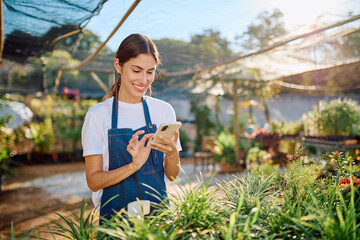Young florist in a sunny garden center uses a smartphone app to manage inventory, customer orders...