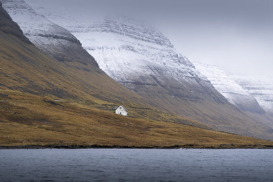 View of a solitary white house nestled at the base of towering, snow-dusted mountains against the moody, grey sky, a serene contrast of color and texture, Torshavn, Faroe Islands.