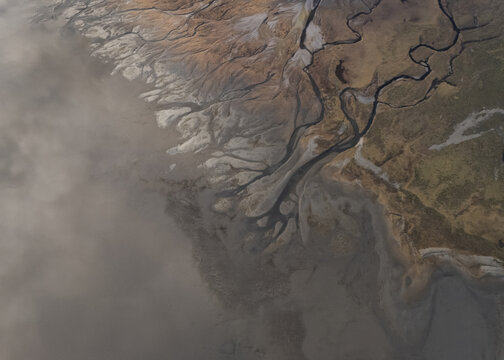 Aerial view of winding rivers carving through the textured earth, juxtaposing the stark land with the soft, enveloping fog, Selfoss, Iceland.