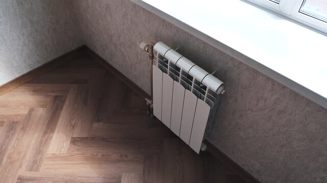 A white bimetallic radiator mounted under a window in a corner of a room with herringbone wooden flooring and patterned wallpaper, creating a cozy corner setup.
