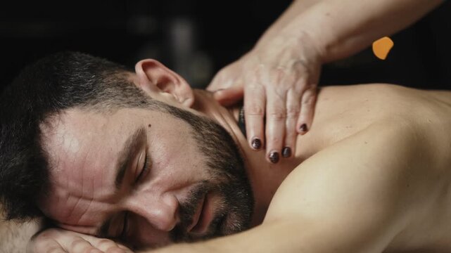 Male client receiving a relaxing hot stone massage in a spa, therapist&rsquo;s hands applying pressure on his back, dim lighting creating a soothing atmosphere. Concept spa, hot stone massage, relaxation, w