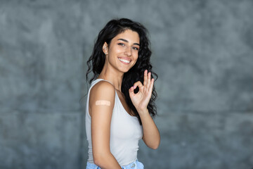 Obraz premium Covid vaccination and healthcare. Cheerful young woman with band aid after coronavirus vaccine injection gesturing okay over grey studio background. Artificial immunity concept