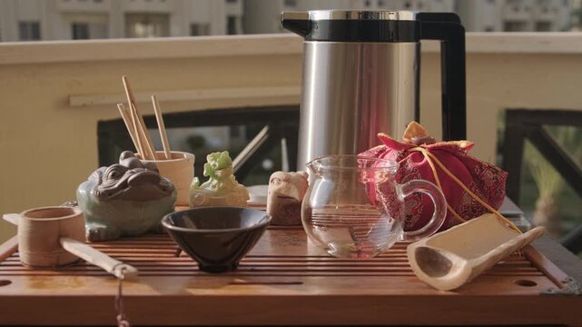Handheld shot of cup, figurines of tea gods, glass pitcher, vacuum flask and bamboo tea strainer at tea tray. Traditional chinese gongfu tea ceremony at home balcony