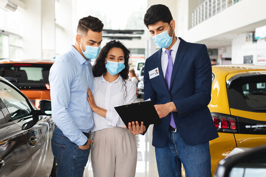 Millennial arab couple in protective face masks visiting auto showroom or exhibition while pandemic, muslim famile embracing while having conversation with salesman, looking at papers in his hands