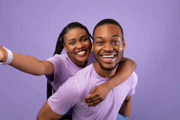 Obraz premium Having Fun. Portrait of happy black couple talking selfie, cheerful woman piggyback riding on her boyfriend's back, holding looking at camera standing isolated over purple studio wall