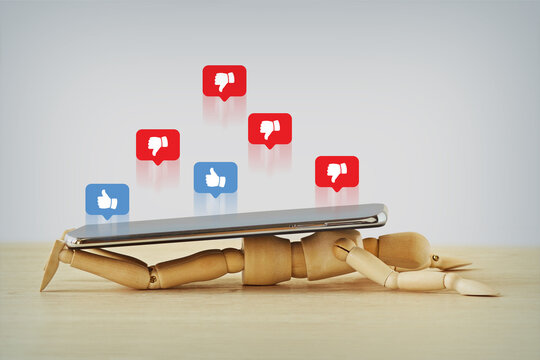 Wooden mannequin crushed under a smartphoe with like and dislike icons in speech bubbles - Concept of Social Media addiction and mental health