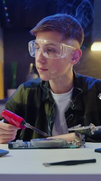 Boy in protective glasses repairs chip and solders printed circuit board with soldering iron. Guy is working on motherboard in evening room. Electronics and soldering concept. Slow motion.