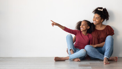 Fototapeta premium Cheerful african american young woman mother and school girl daughter in similar outfits sitting on floor at home, hugging, smiling, teen girl pointing at copy space for advertisement, panorama
