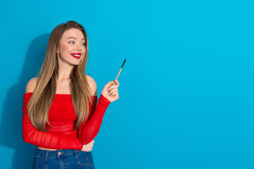 Obraz premium Beautiful young woman holding a pen on a blue background, smiling while wearing a red stylish top and denim jeans