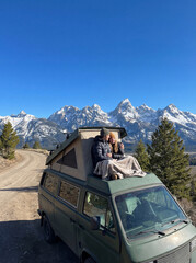 A young couple in love sit on a camper, admiring the view of the mountains and freedom