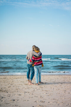 ture couple standing on the beach with arms around, looking at the sea