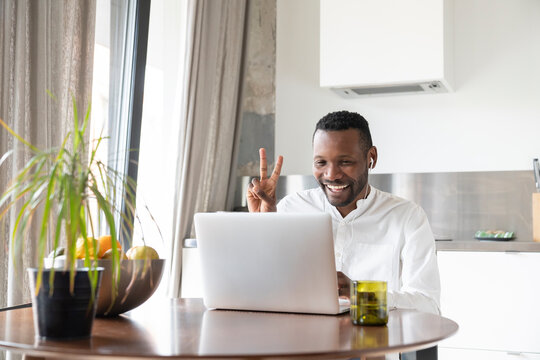 Portrait of happy man sitting at kitchen table during video chat showing victory sign