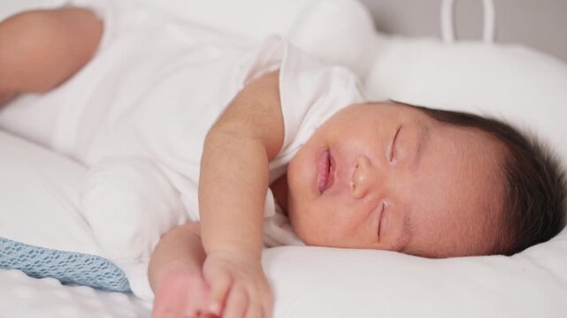 Adorable Asian newborn baby smiling in peaceful sleep on soft white pillow, symbolizing love, care, and comfort.