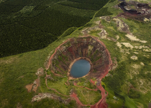 Aerial view of the Kerid Crater Lake with its vibrant turquoise waters nestled within striking red volcanic slopes, surrounded by lush green vegetation, Flugir, Rangarthing ytra, Iceland.
