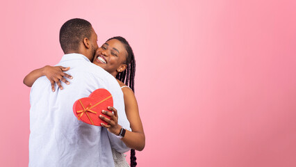 Passionate black lovers hugging on pink background, lady holding heart shaped gift box....