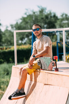 Smiling young man with earbuds and longboard sitting on top of halfpipe in skatepark