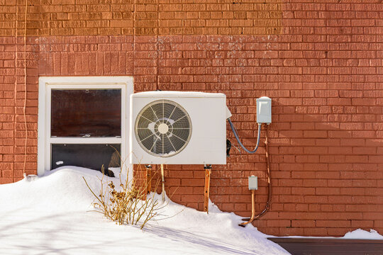 Wall-mounted ductless heat pump compressor  on the exterior of a red brick residential building on a snowy winters day.