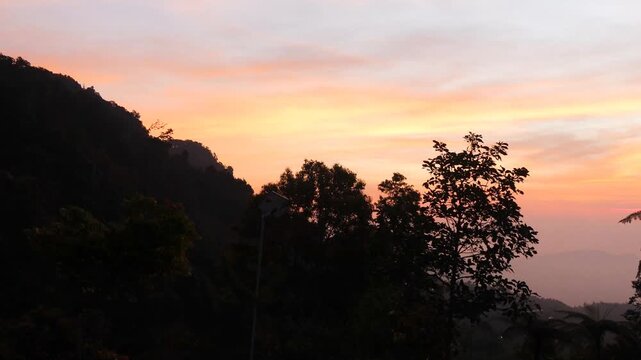 A beautiful sunrise with bright orange and pink hues over the silhouettes of windblown trees and tropical mountains, creating a peaceful and dramatic natural morning scene.
