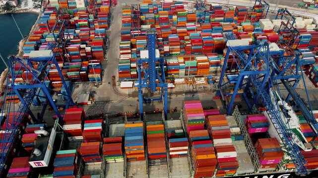 Top-down aerial drone view of port cranes loading and unloading shipping containers at a busy cargo terminal. Industrial logistics, global trade, maritime transport and supply chain infrastructure con