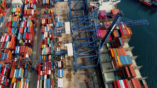 Top-down aerial drone view of port cranes loading and unloading shipping containers at a busy cargo terminal. Industrial logistics, global trade, maritime transport and supply chain infrastructure con