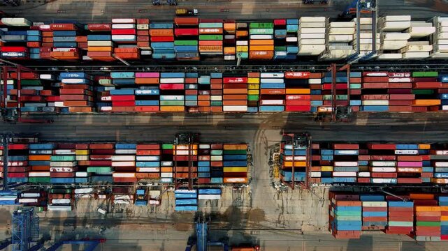 Top-down aerial drone view of a busy container port terminal with stacked shipping containers, cargo ship and cranes. Global trade, logistics, maritime transport and industrial infrastructure concept.