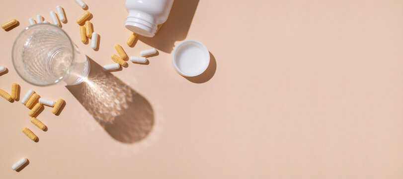 Glass of water, white and beige vitamin capsules, supplements, or tablets on beige background, flat lay top view. Minimalistic medical background. Sunny shadows. Dietary supplements, health care