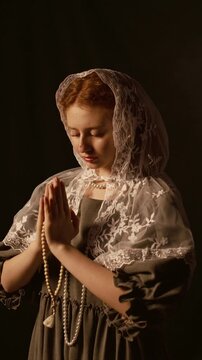 Vertical dolly in shot of teenage ginger haired girl wearing white lace chapel veil clasping hands with rosary while praying under low key light in studio against back background, slow motion
