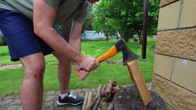 A man uses an axe to split firewood on a log outdoors, preparing fuel for grilling or a campfire in a garden setting.